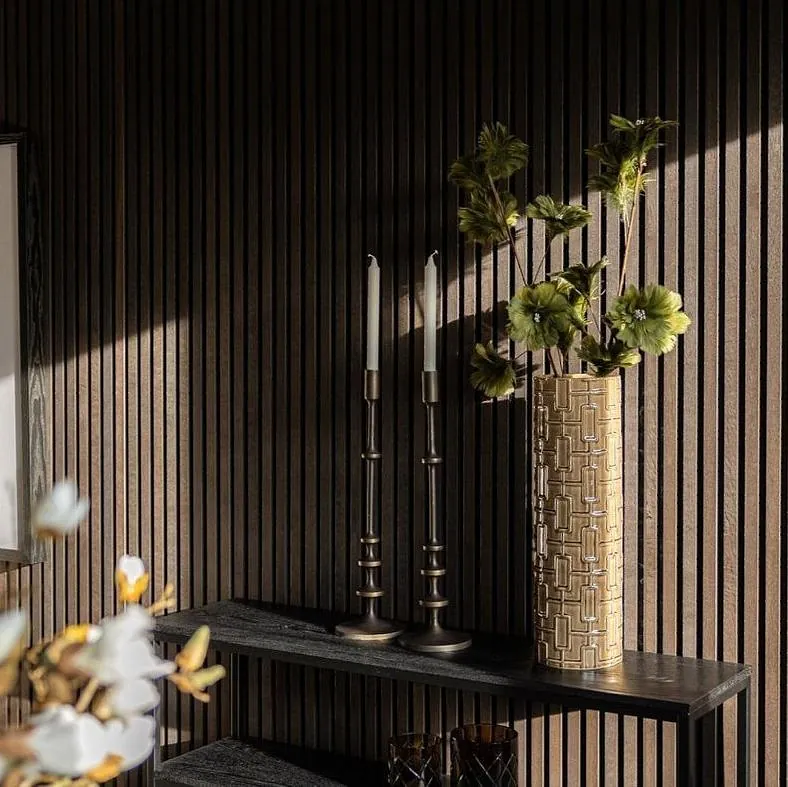 Mørk trevegg med dekorhyller, høye lysestaker, blomster i gullvase og kunstbilde.